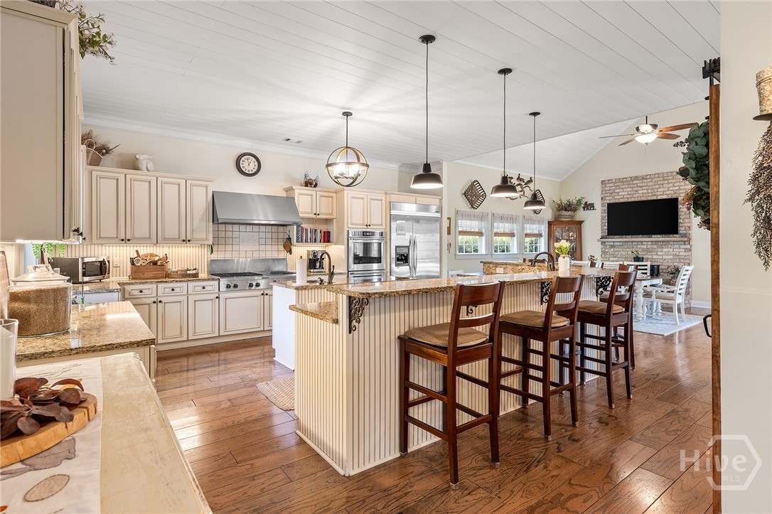 The main kitchen with two granite islands, cream raised-panel cabinetry, a stainless gas range, pendant lighting, and a vaulted shiplap ceiling, open to the family room with a brick fireplace