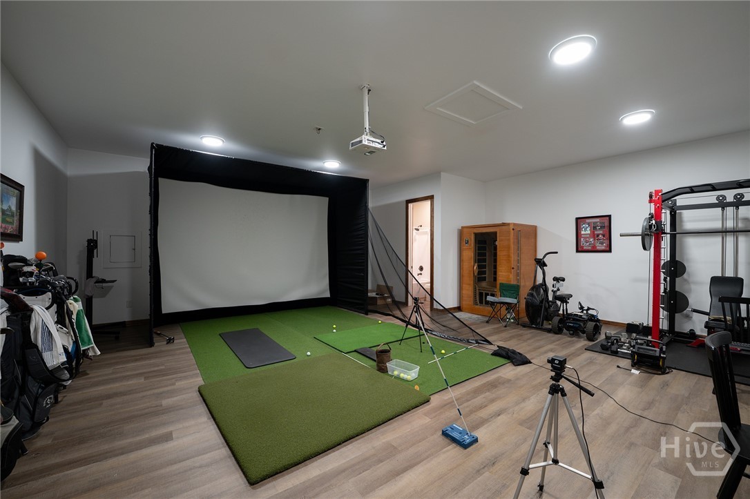 The detached shop interior outfitted as a recreation room with a golf simulator screen and putting turf, a home gym with a cable machine and exercise bike, and an infrared sauna along one wall