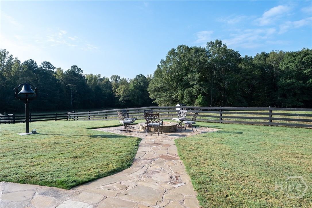 A flagstone fire ring with a circle of patio chairs and a mounted dinner bell, set on the lawn and overlooking four-board-fenced horse pastures and the tree line beyond